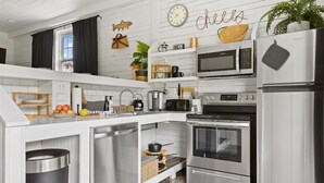 Una cocina moderna con armarios blancos, electrodomésticos de acero inoxidable, microondas, horno y refrigerador. Hay estantes con objetos decorativos y un reloj en la pared.