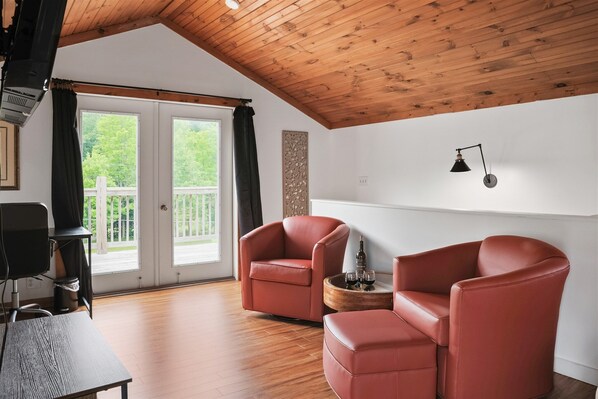Une pièce avec un plafond en bois, une porte-fenêtre coulissante donnant sur un balcon, deux fauteuils en cuir rouge, une petite table en bois avec une bouteille et des verres, et une lampe de bureau noire.