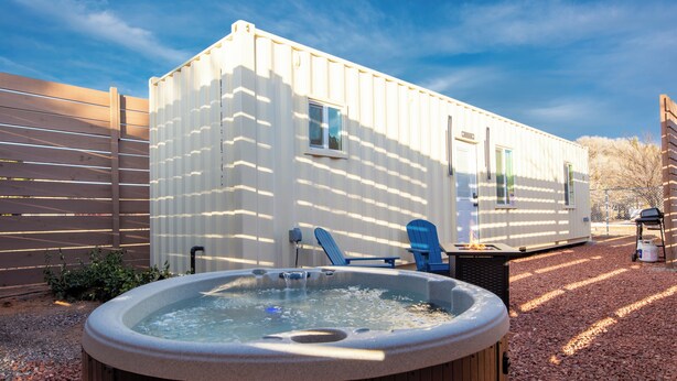 Un bain à remous avec une terrasse en bois, une chaise bleue et une structure mobile de type caravane, dotée de fenêtres et d’une porte.