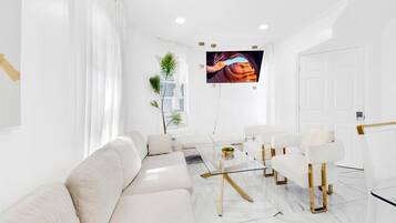 Un salon moderne avec un canapé, une table basse et des fauteuils, un téléviseur à écran plat fixé au mur, et une porte sur le côté droit.