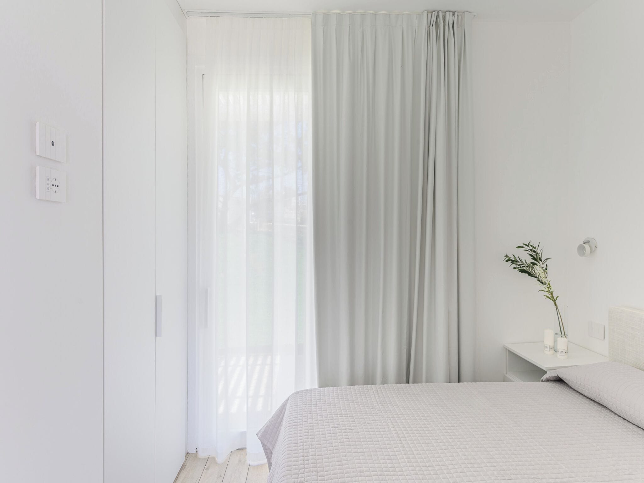 Egy letisztult, minimalista hálószoba, amelyben egy ágy, egy éjjeliszemélyzet, egy növényt tartalmazó váza és könnyű, áttetsző függönyök találhatók.