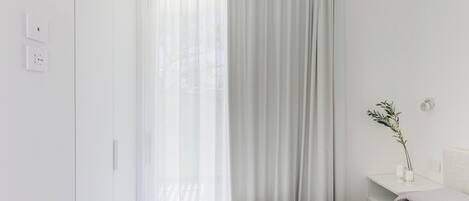 A minimalist bedroom with a bed, a nightstand, a vase with a plant, and sheer curtains.
