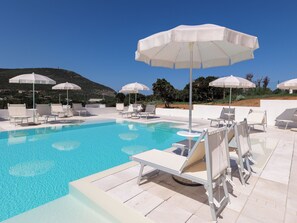 Zona de piscina con sillas reclinables blancas y sombrillas, rodeada por un muro blanco y vistas a un paisaje montañoso.