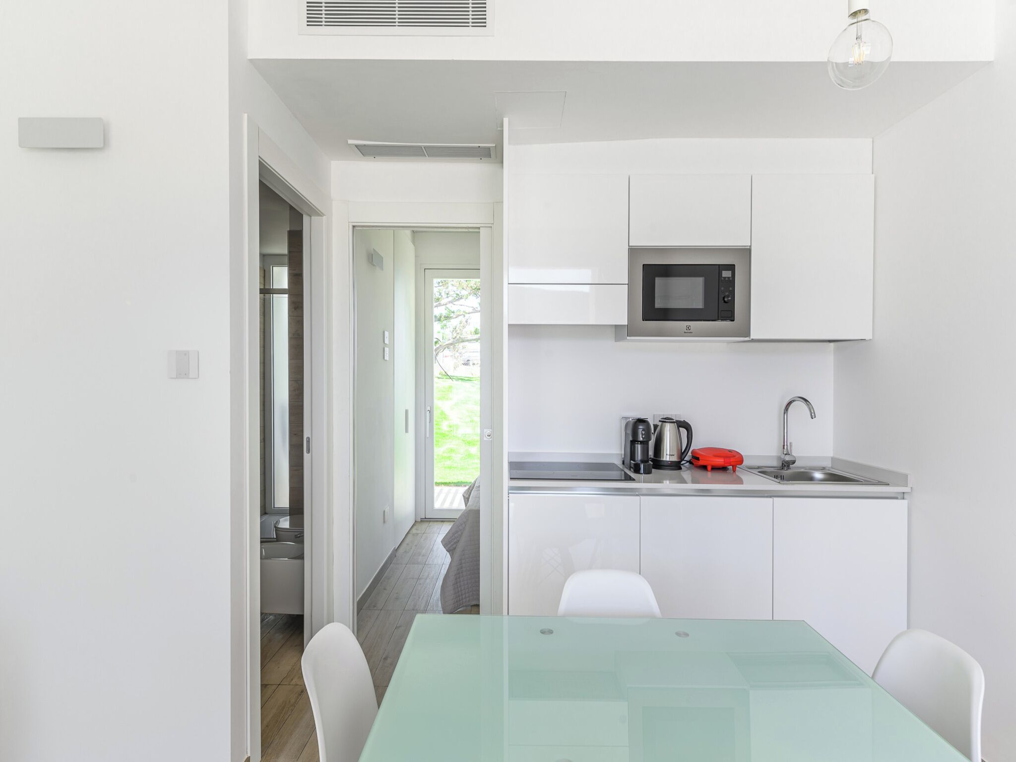 A compact kitchen with white cabinets, a microwave, a kettle, and a sink. There is a glass table with chairs.