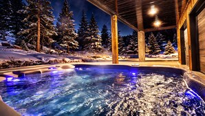 Un jacuzzi con terraza de madera y vistas a árboles nevados en la noche.