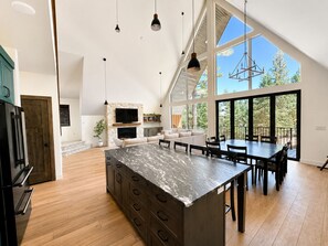 A spacious kitchen with a large island, a dining area with a table and chairs, a built-in fireplace, and large windows offering a view of the outdoors. - Modern Mountain A-Frame w/ Hot Tub + Sauna + Fire Pit - Sleeps 10 -Pet Friendly (Duck Creek Village)