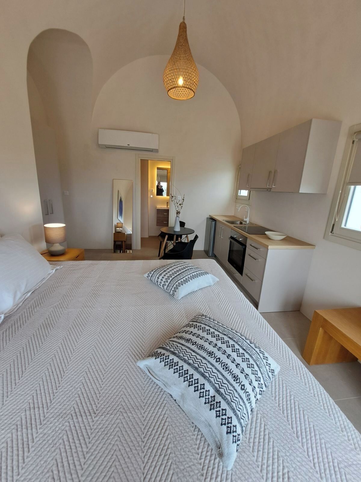 A modern bedroom with a large bed, a hanging light fixture, and a view into the kitchen.