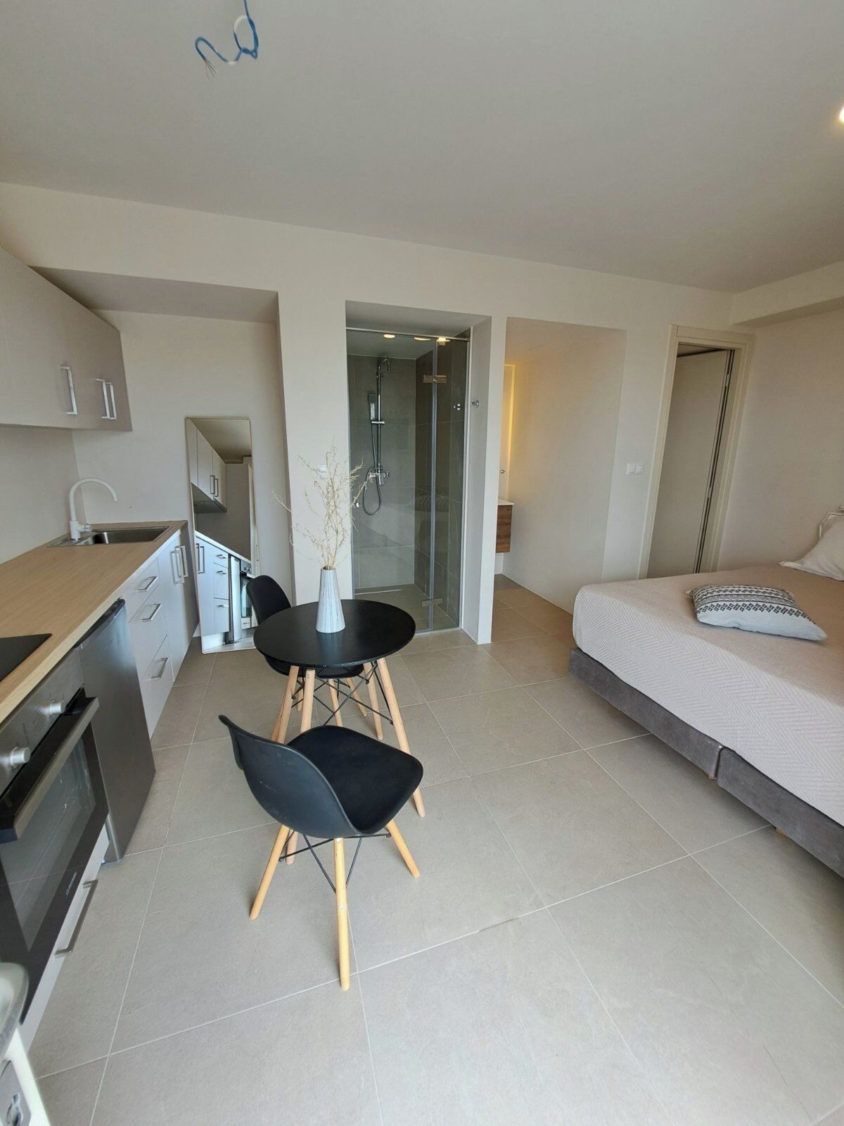 A modern hotel room with a kitchenette, a dining area with a table and chairs, a shower area, and a bed.