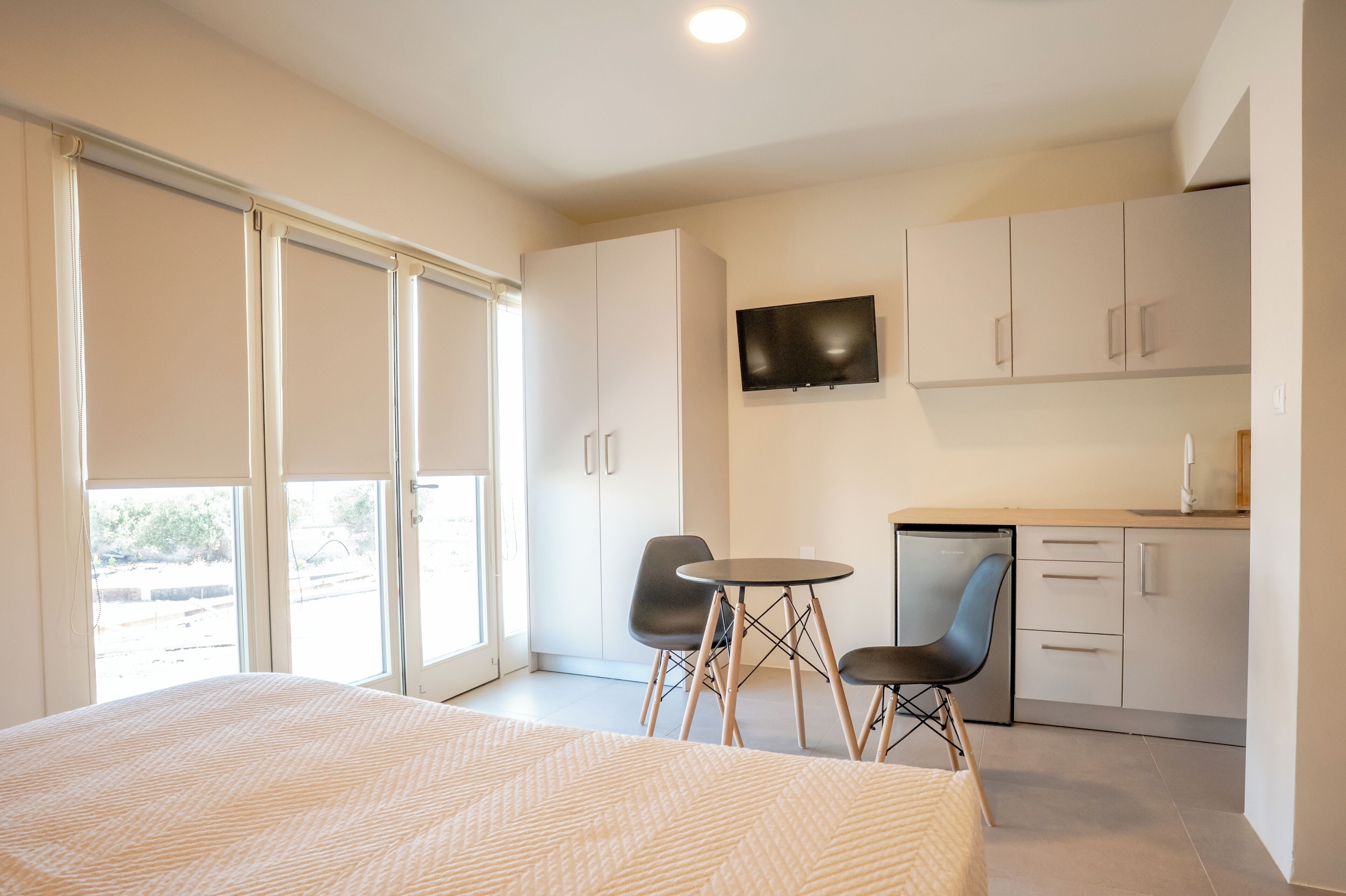 A modern bedroom with a large bed, a small dining table with two chairs, a kitchen area with cabinets, and a TV mounted on the wall.