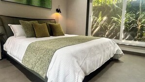 Une chambre avec un grand lit, une table de chevet, une lampe fixée au mur et une fenêtre donnant sur un paysage verdoyant.