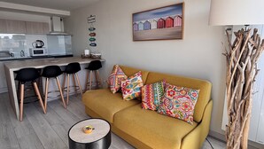 Een moderne woonkamer met een mosterdgele bank, kleurrijke kussens, een ronde salontafel en een hoge houten sculptuur.