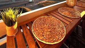 Een balkon met geweven matten, een potplant en uitzicht op de ondergaande zon boven een kustweg.
