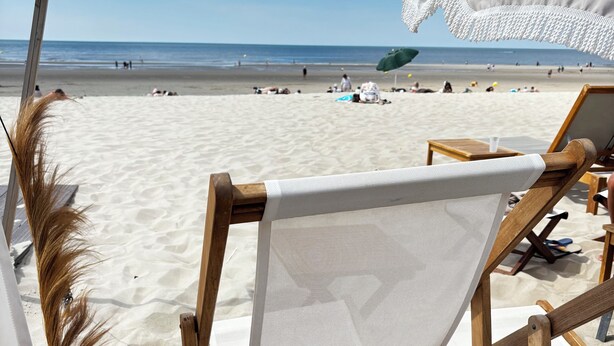 Een strandstoel met een witte mesh rugleuning en houten frame, geplaatst op zand met een helder uitzicht op het strand en de oceaan.