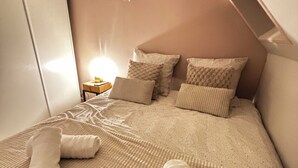 Une chambre avec un plafond en pente, un lit avec une literie beige, plusieurs oreillers et une table de chevet avec une lampe.
