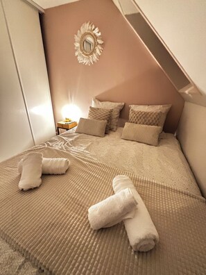 Une chambre avec un plafond en pente, un lit avec une literie beige, plusieurs oreillers et une table de chevet avec une lampe.
