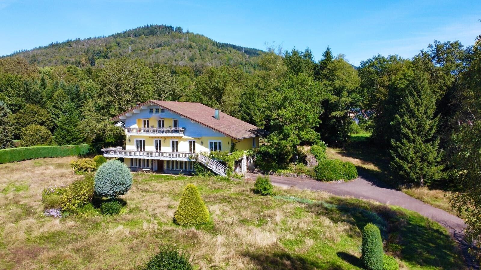 Uma casa de dois andares com varanda, cercada por um gramado bem cuidado e árvores.