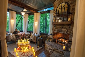 A cozy room with a stone fireplace, a seating area with a coffee table adorned with candles and a romantic message, and large windows offering a view of trees. - Stonara • Lovers’Hideaway: Cliffside Hot Tub + Sauna + Cinema (Blue Ridge)
