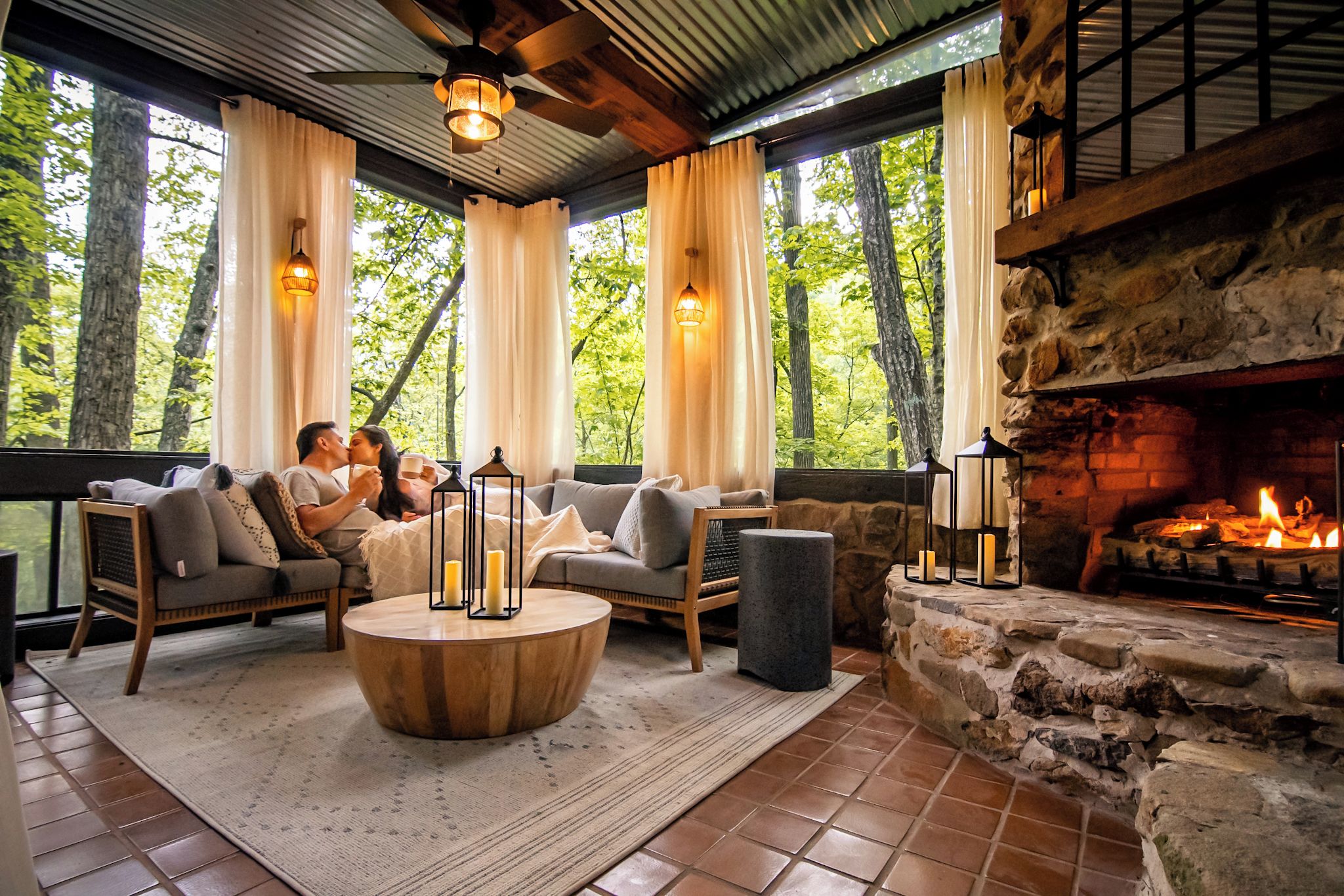 Un coin salon confortable avec une cheminée en pierre, de grandes fenêtres avec des rideaux et un ventilateur de plafond.