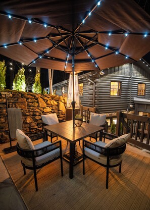 Un área de comedor al aire libre con una mesa y sillas, un muro de piedra y una cabaña de madera al fondo.