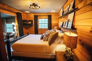 Interior de cabana de madeira com cama, televisão na parede e janela com cortinas.