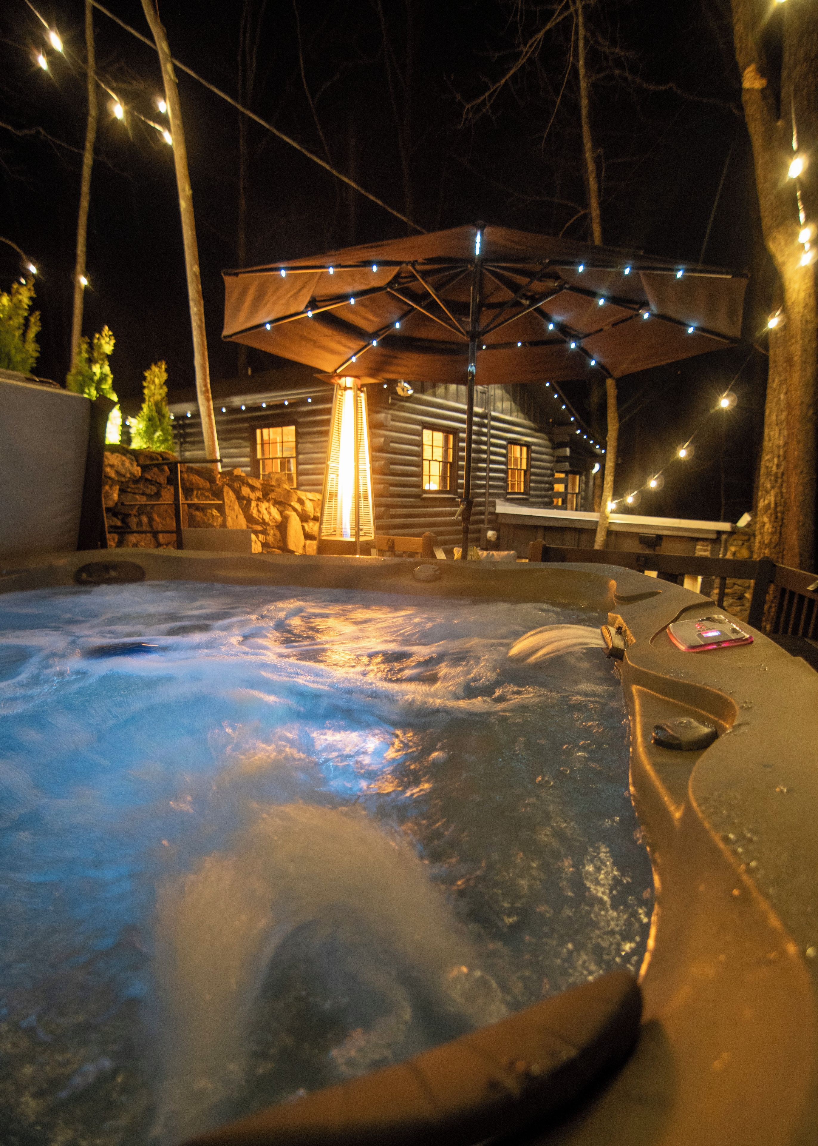 A hot tub with a wooden deck, surrounded by a stone wall and a wooden cabin in the background.
