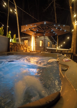 Uma banheira de hidromassagem com deck de madeira, rodeada por um muro de pedra e uma cabana de madeira ao fundo.