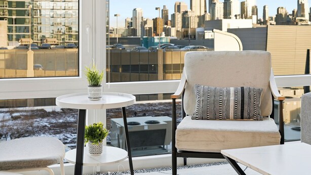 A modern balcony with a city view, a cushioned chair, a small table with a plant, and a potted plant on the floor.