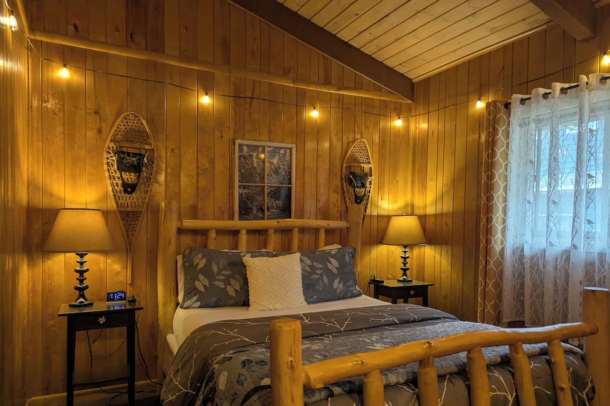A wooden cabin bedroom with a bed, two bedside lamps, a clock, and a window with curtains.