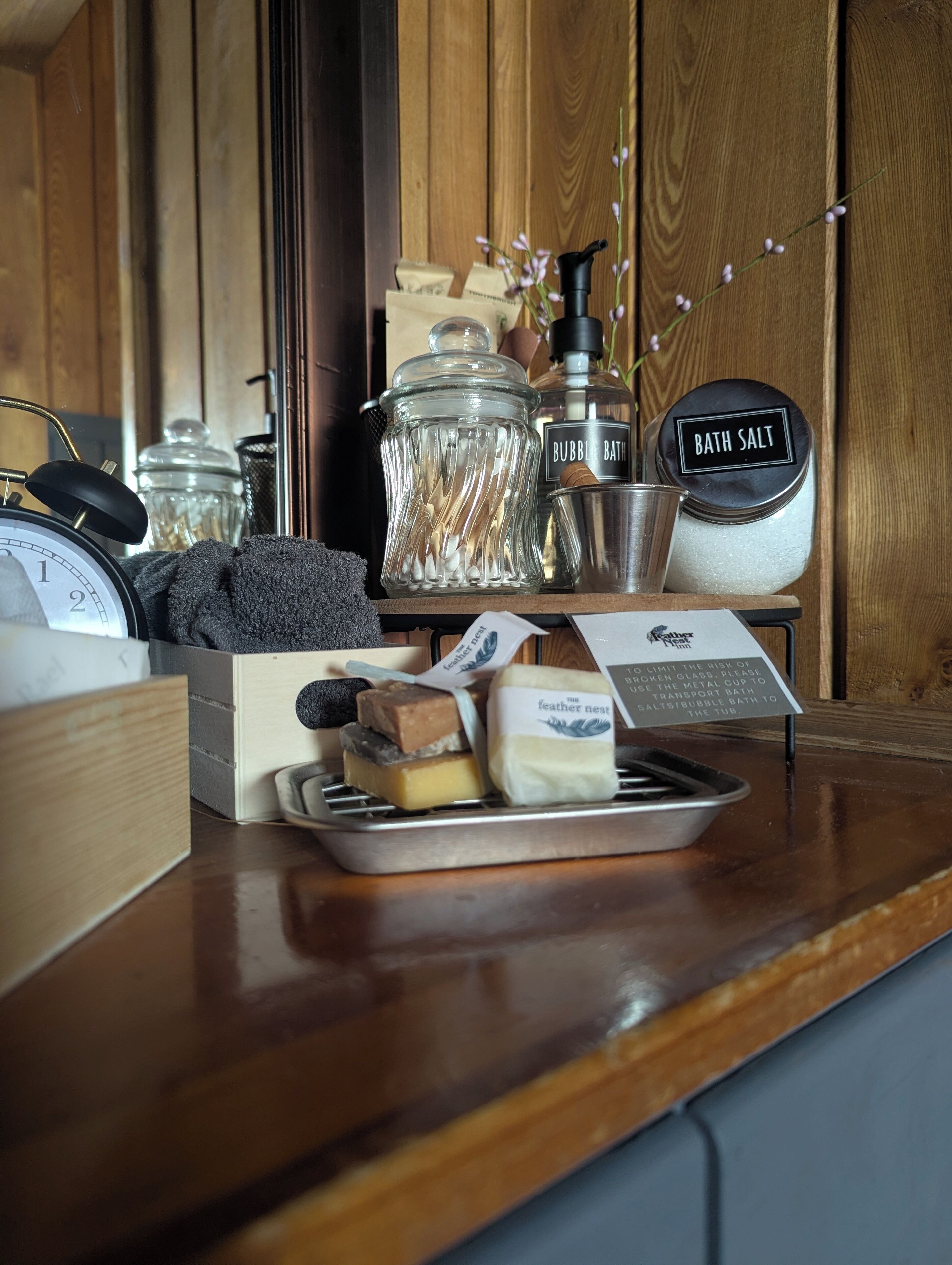 Una encimera de madera con una taza de metal, un tarro de vidrio con la etiqueta 'BUBBLE BATH', un recipiente metálico etiquetado 'BATH SALT' y una bandeja con jabón y una pastilla de jabón con plumas.