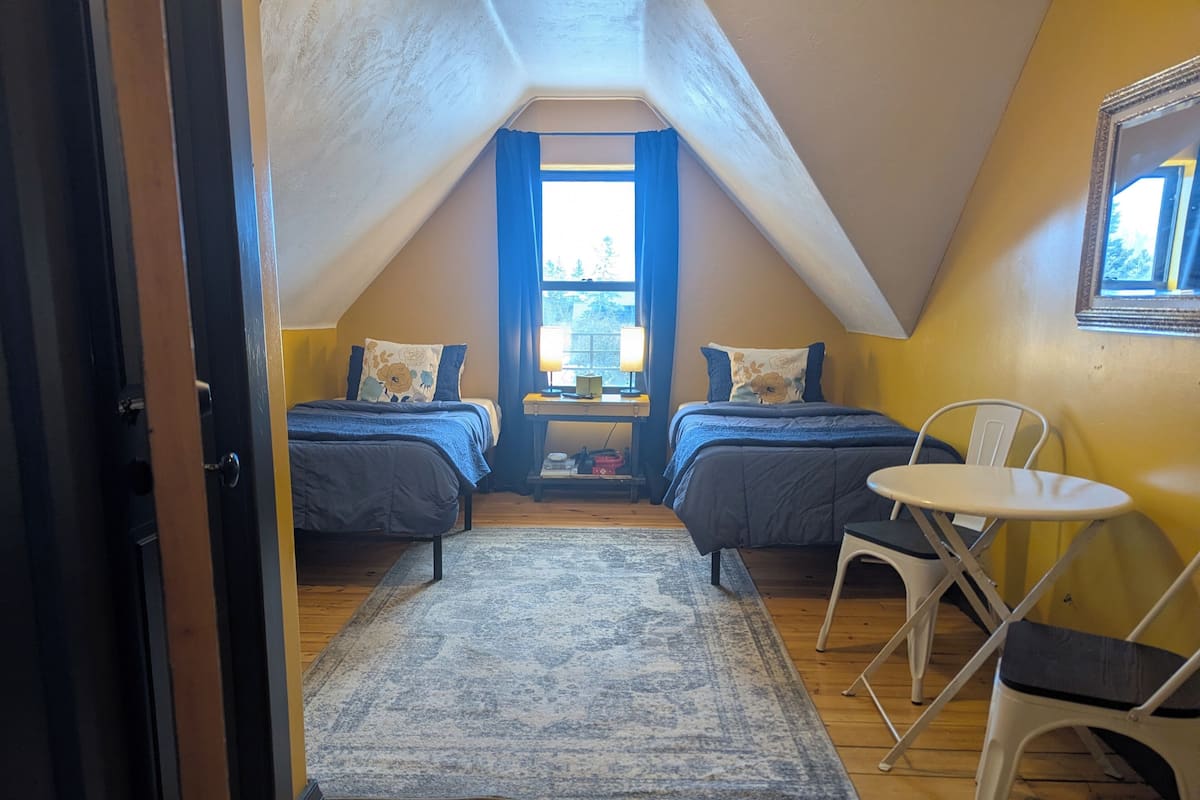 A small attic bedroom with two beds, a desk, a chair, and a window.
