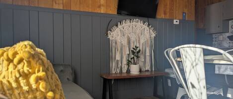 A cozy room with a grey sofa, a wooden accent wall, a macrame wall hanging, a small table with a potted plant, and a white chair.
