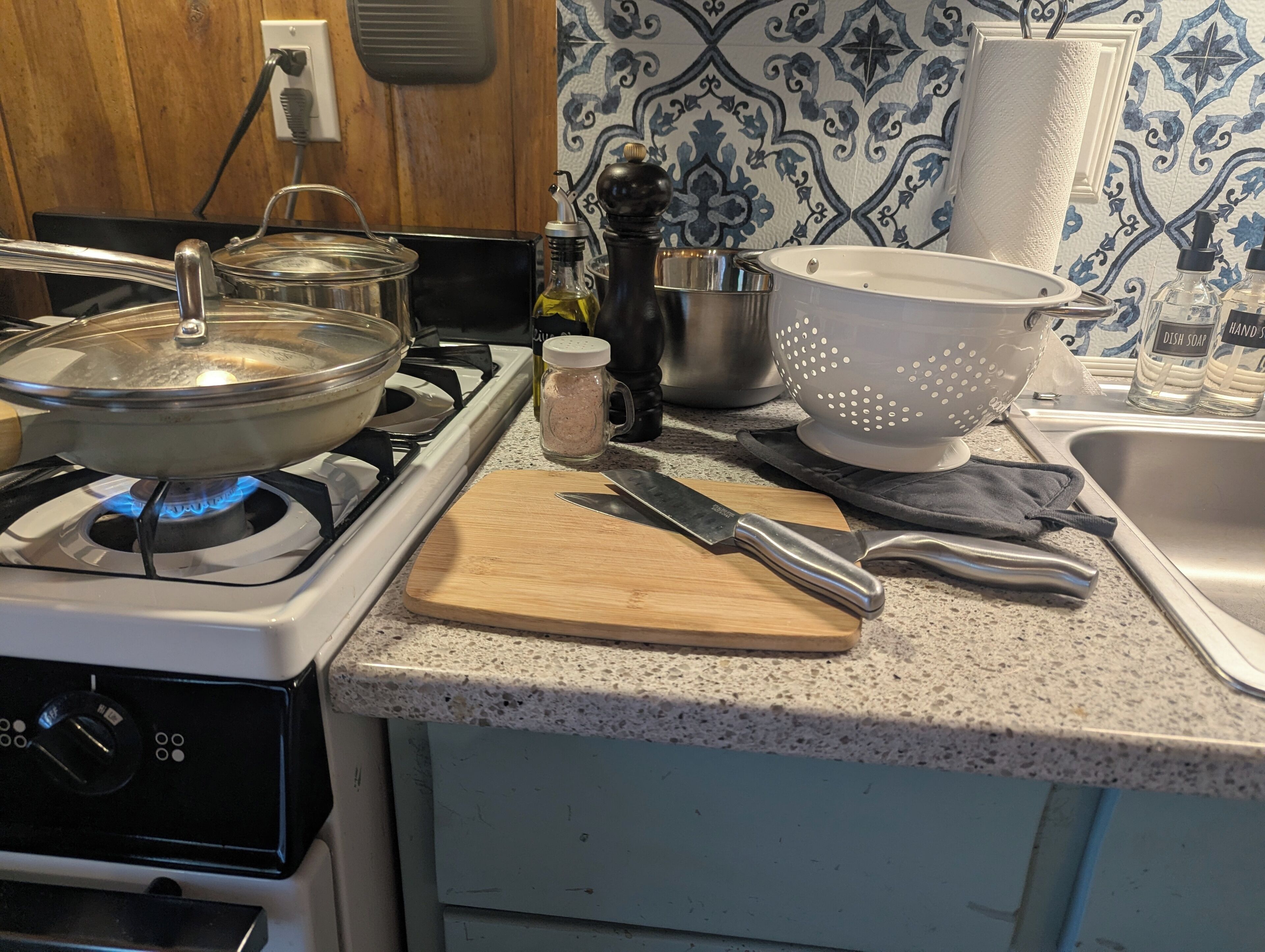 A kitchen with a gas stove, a cutting board, a knife, a colander, and a sink.