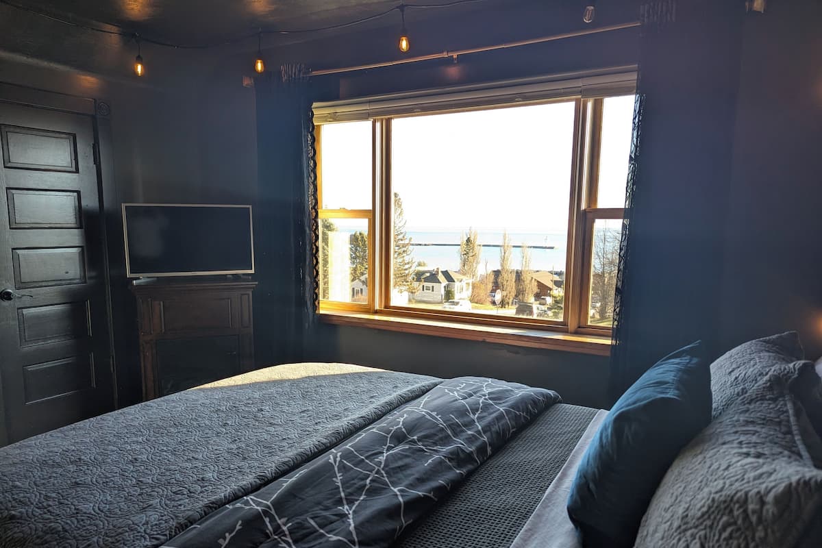 A bedroom with a bed, a TV, a window with a view of houses and trees, and a door.
