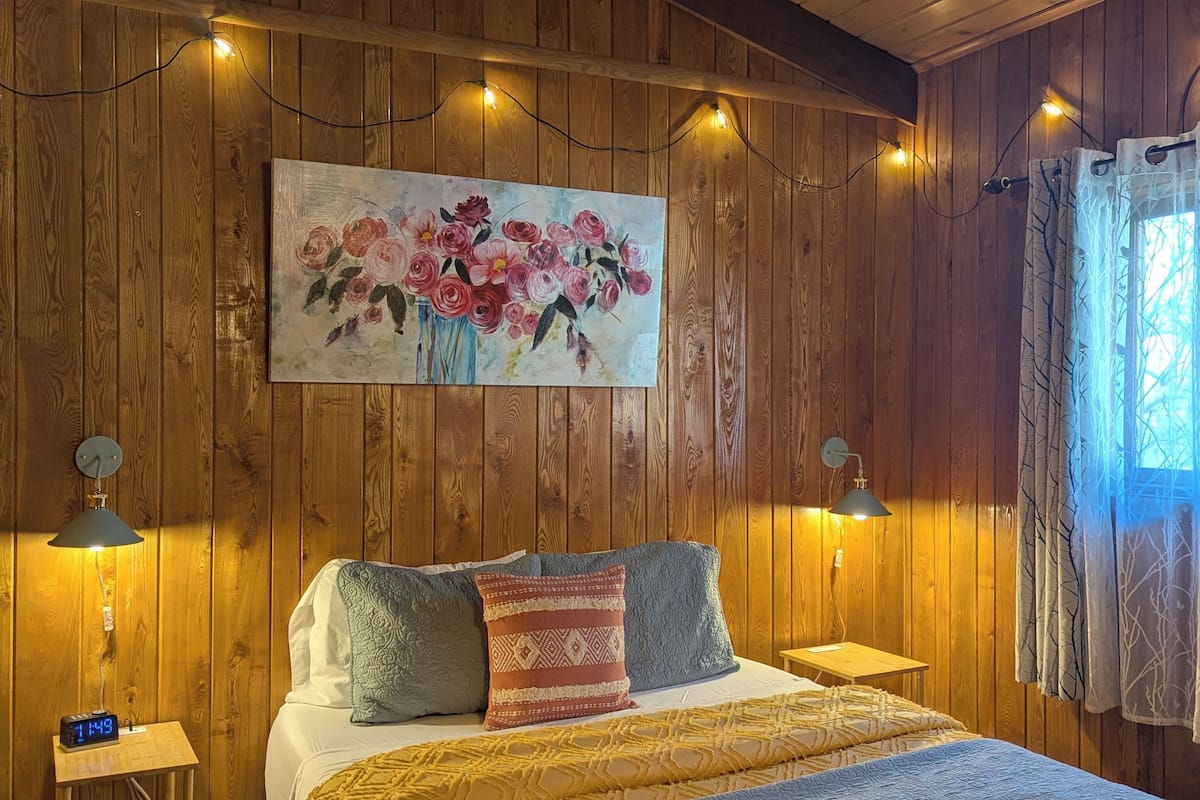 A wooden-paneled bedroom with a bed, two bedside lamps, a painting of flowers, and a clock on a nightstand.