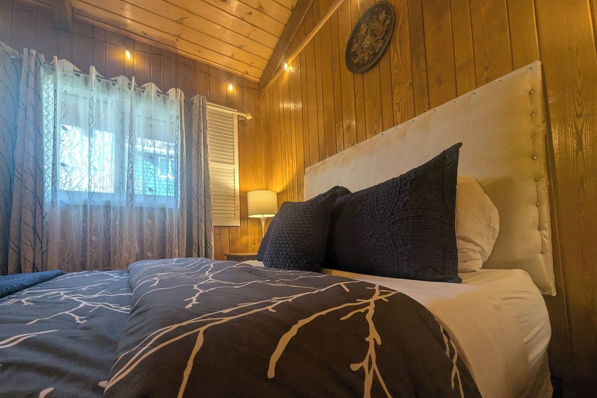 A wooden-paneled room with a bed, a nightstand, a lamp, and a window with curtains.