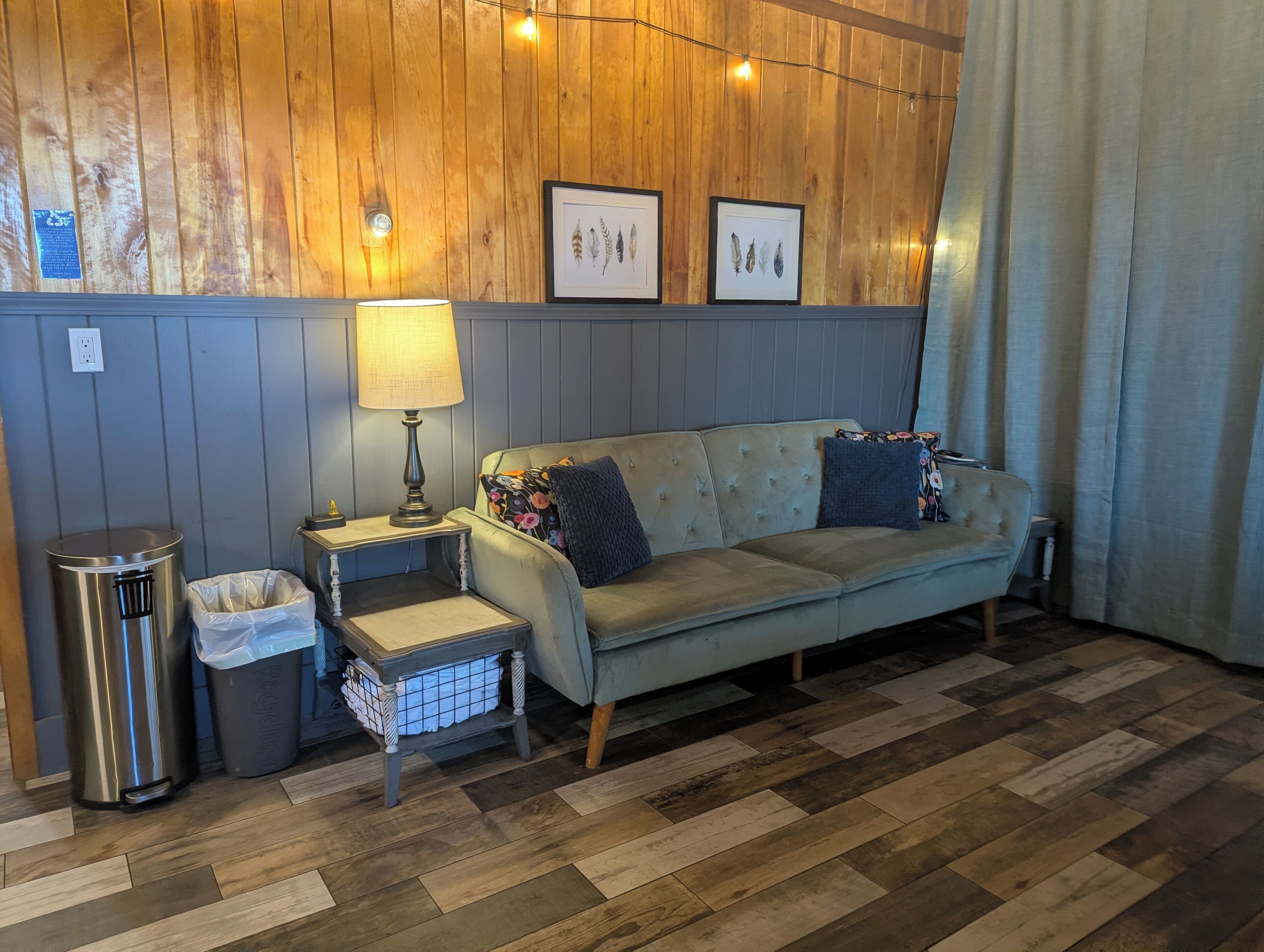 A room with a wooden paneled wall, a grey sofa with floral cushions, a small table with a lamp, a trash bin, and a picture frame.