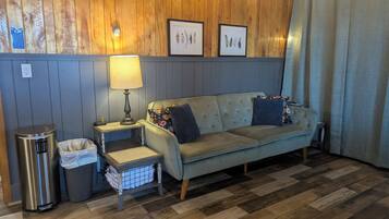 A room with a wooden paneled wall, a grey sofa with floral cushions, a small table with a lamp, a trash bin, and a picture frame.