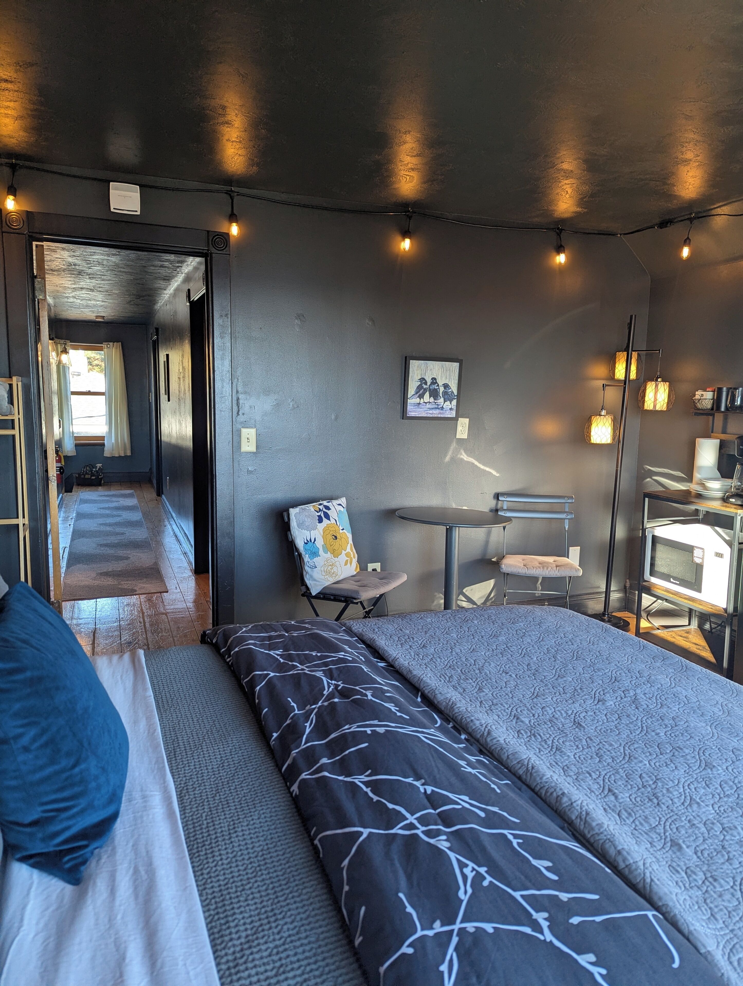 Una habitación moderna con cama, escritorio y silla. Tiene una ventana con cortinas, una mesita pequeña y una lámpara.