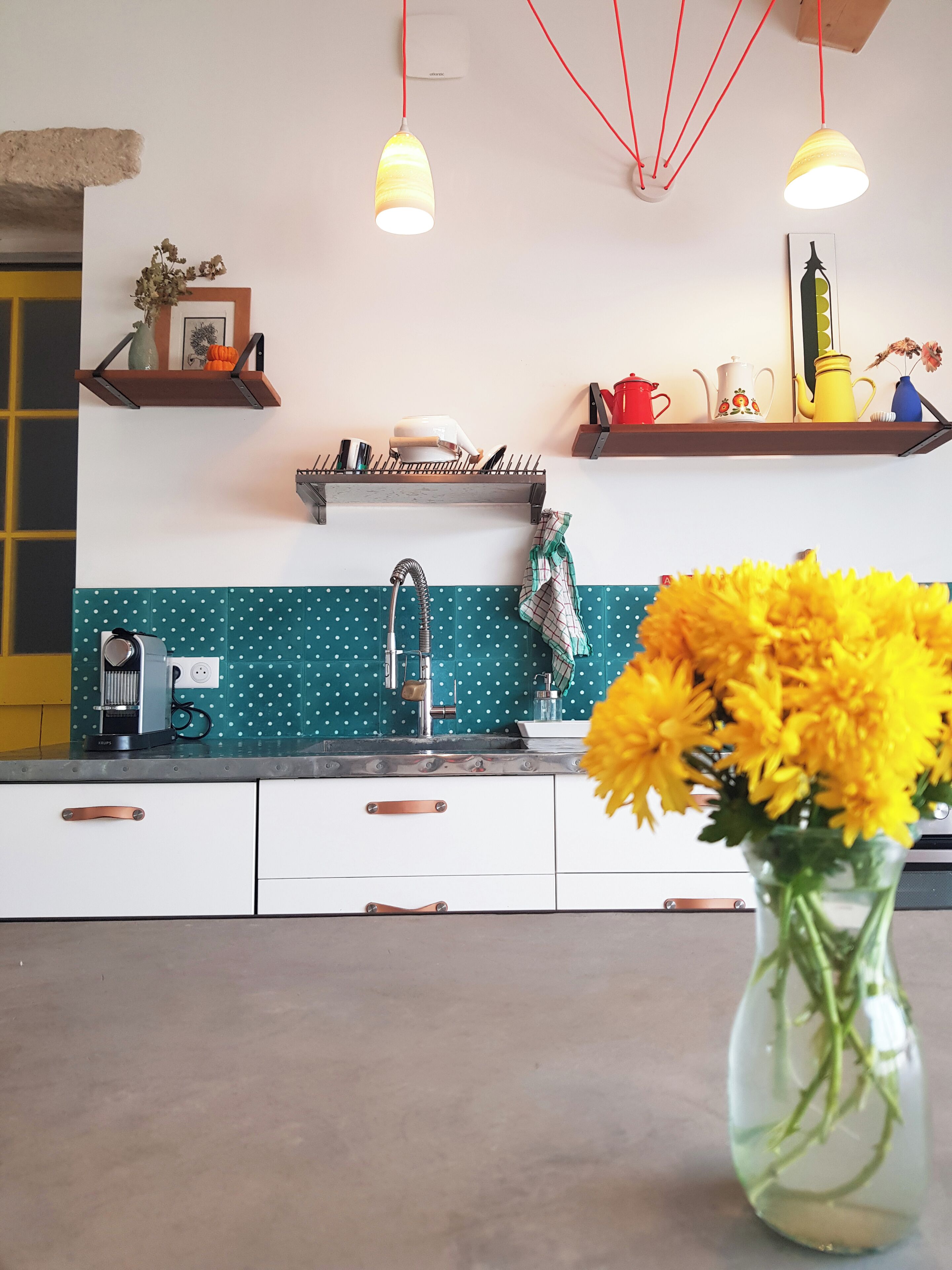 Una cocina con salpicadero color turquesa, armarios blancos y una cafetera. Hay dos lámparas colgantes y un jarrón con flores amarillas en la encimera.