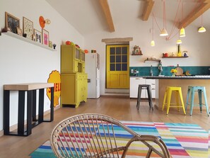 A kitchen with a yellow door, a white refrigerator, and a wooden table. There are colorful stools and a wicker chair. - Unique House in the Luberon with an Above-Ground Pool   (Robion)