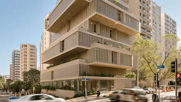A multi-story building with a modern architectural design, featuring balconies with greenery, a street view with cars and a pedestrian crossing, and a clear sky.