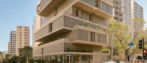 Un edificio de varios pisos con un diseño arquitectónico moderno, que cuenta con balcones con vegetación, vista a la calle con coches y un paso de peatones, y un cielo despejado.
