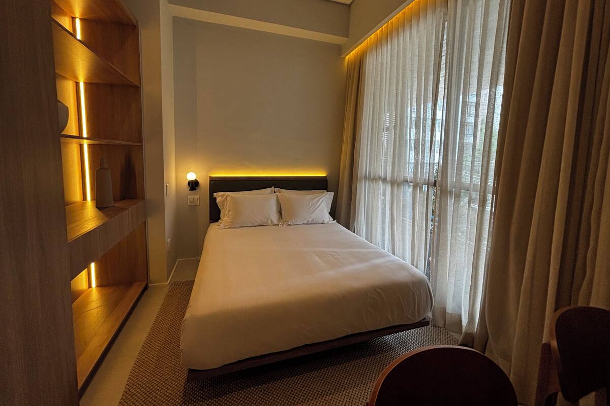 A hotel room with a bed, a nightstand, a lamp, a window with curtains, and a wooden shelf.