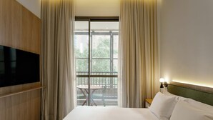 A hotel room with a bed, a TV mounted on the wall, a window with curtains, and a small table with chairs outside. - Aloe Casa Jardins (São Paulo)