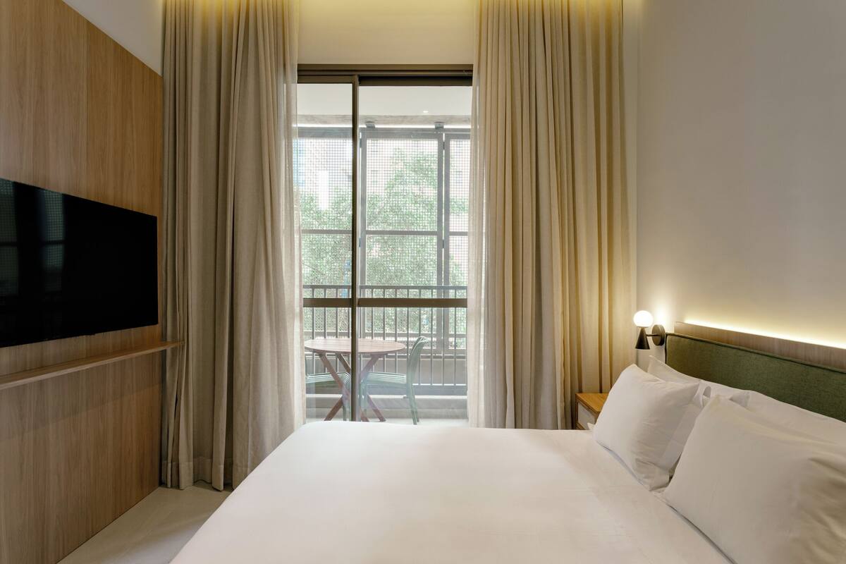 A hotel room with a bed, a TV mounted on the wall, a window with curtains, and a small table with chairs outside.
