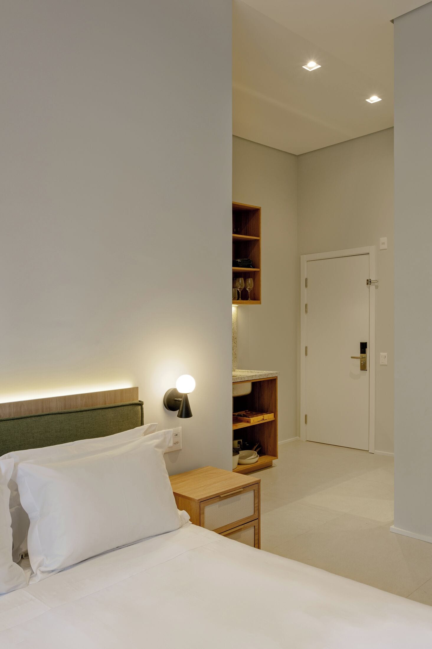 Habitación de hotel con cama, mesita de noche, lámpara de pared y una puerta que da a otra habitación.