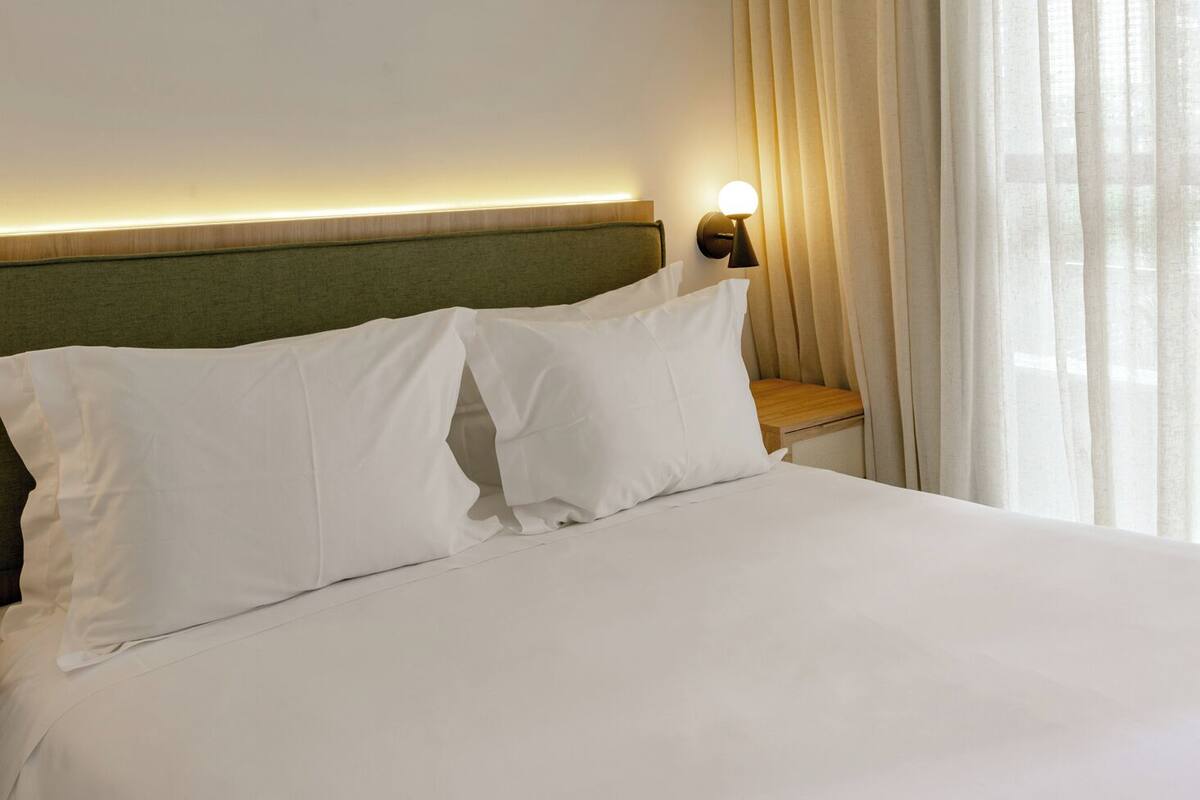 A hotel room with a bed, a nightstand with a lamp, and sheer curtains.