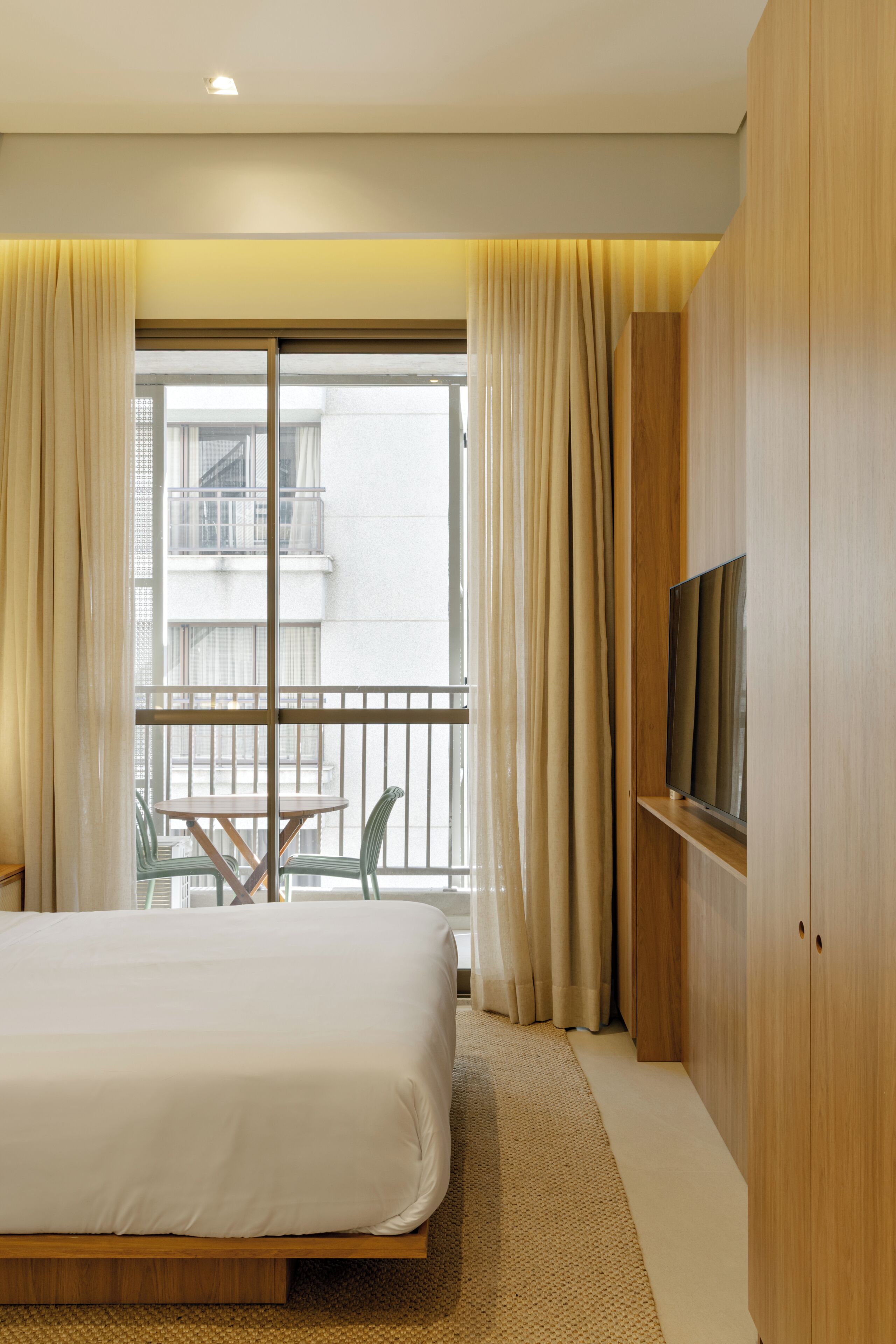 Habitación de hotel con cama, televisor montado en la pared, balcón con mesa y sillas, y cortinas.