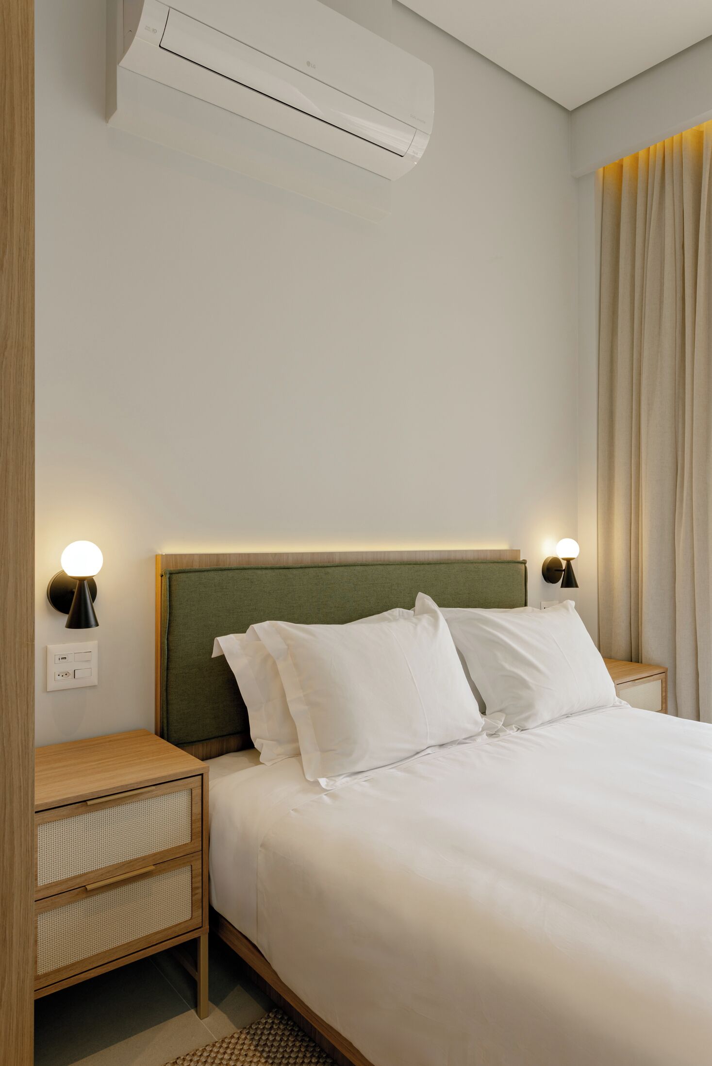 A hotel room with a bed, two bedside tables, an air conditioner, and a wall-mounted light fixture.