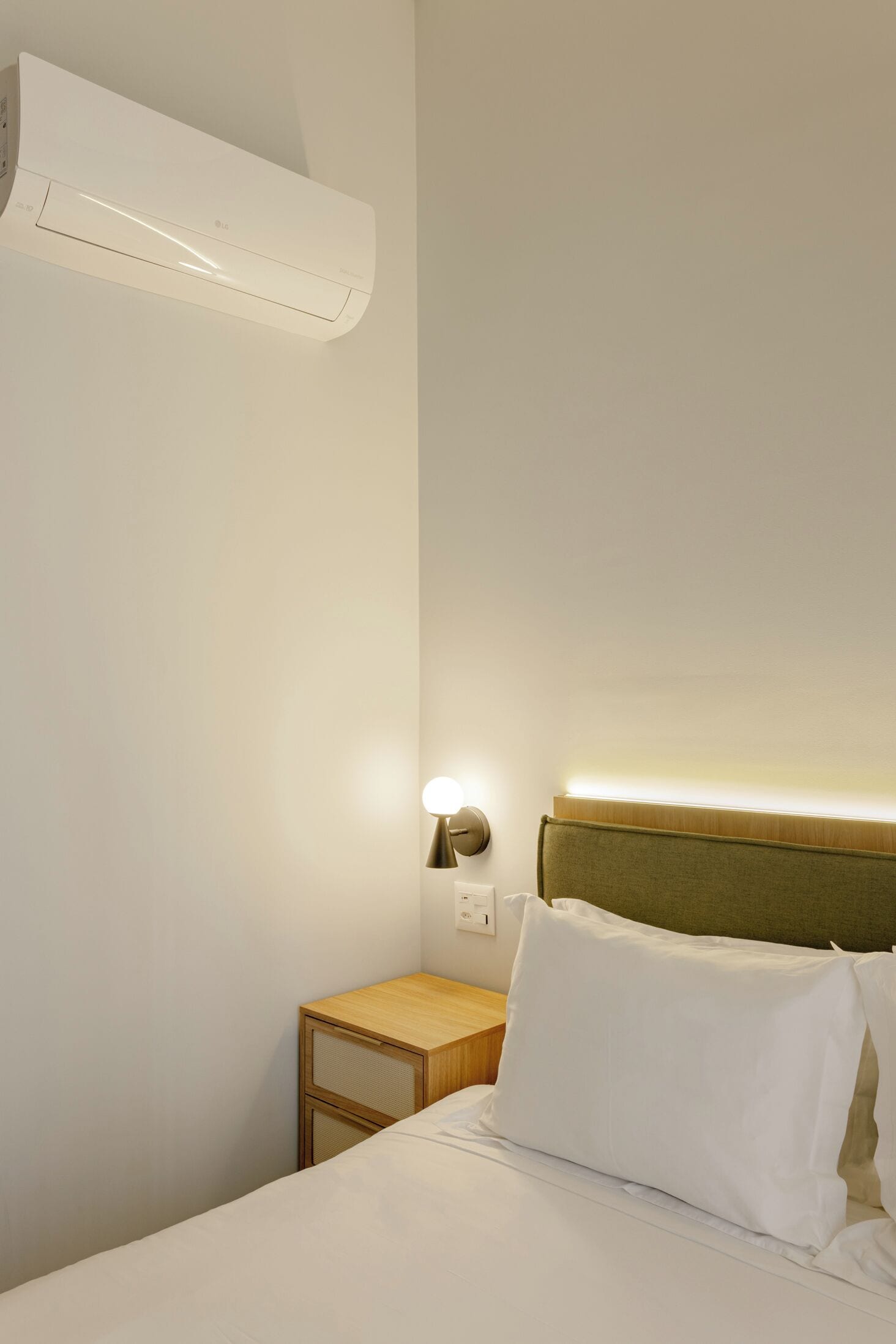 A hotel room with a bed, a bedside table, a lamp, and an air conditioning unit.